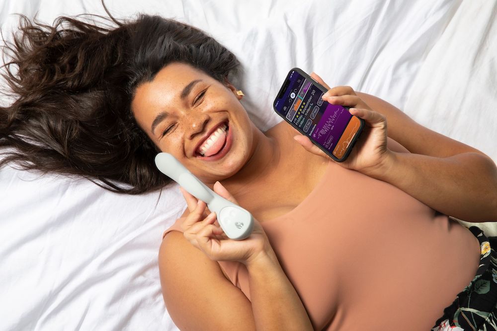 woman with brown hair laying in bed sticking tongue out holding grantie grey lioness smart vibrator and lioness health app