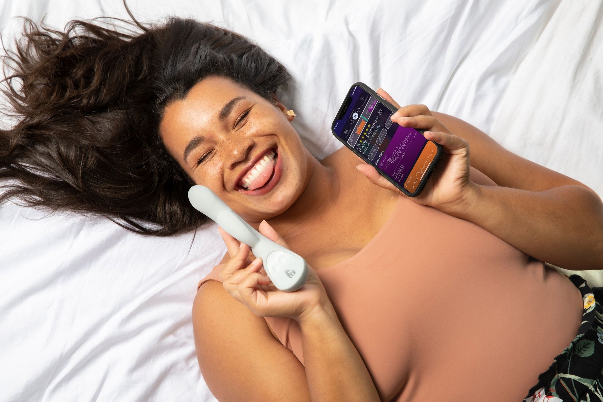 Woman laying on a bed and sticking out her tongue while holding Lioness Smart Vibrator and App